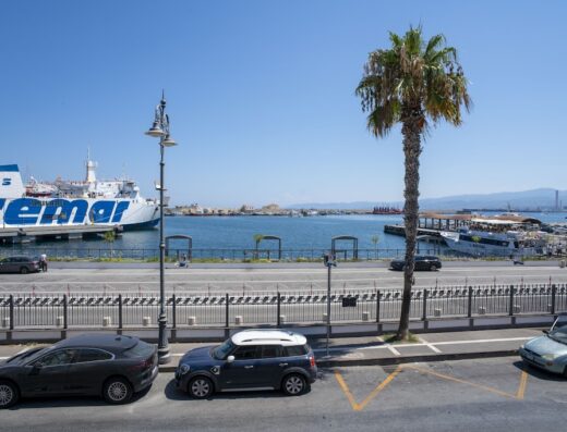 Immagine dell'Hotel Petit con vista sul porto di Messina, Sicilia. Si vedono traghetti, auto parcheggiate, una palma e il lungomare.