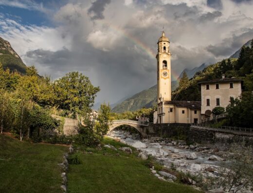 Paesaggio pittoresco a Piuro, Italia, con un arcobaleno che si estende su una chiesa storica e un fiume roccioso, circondato da montagne verdi.