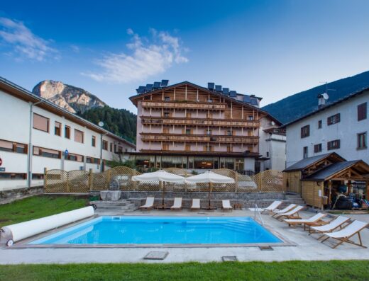 Hotel Posta: Piscina esterna con lettini e ombrelloni, circondata da edifici e montagne, ideale per il relax e il turismo.