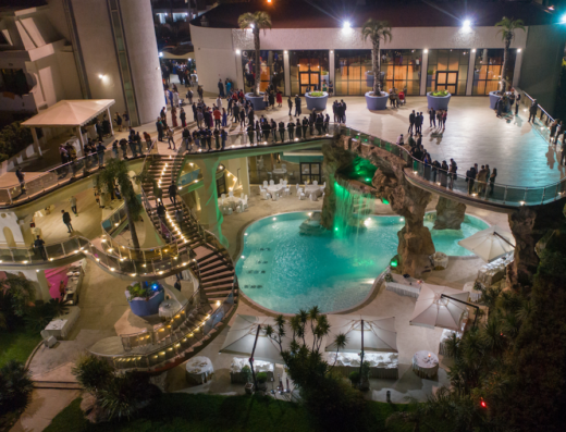 Vista aerea notturna dell'Hotel Residence Arcobaleno con piscina illuminata, cascata artificiale, scalinata a spirale e ospiti su passerella.