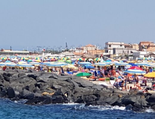 Affollata spiaggia di Fiumicino con ombrelloni colorati e persone che prendono il sole, vista dal molo roccioso.