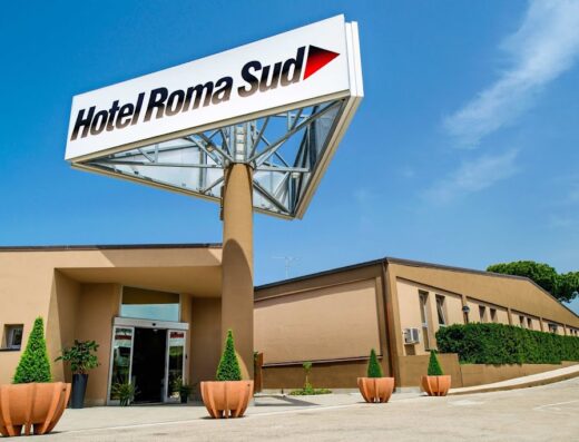 Hotel Roma Sud entrance with large sign and automatic doors, landscaping with potted plants, clear blue sky.