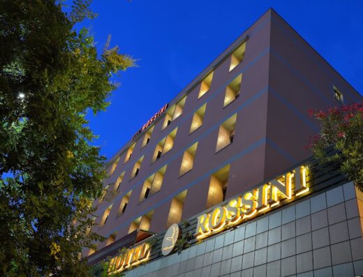 Esterno dell'Hotel Rossini al crepuscolo, con insegna luminosa "Rossini", finestre illuminate e vegetazione circostante. Hotel di lusso e accoglienza.