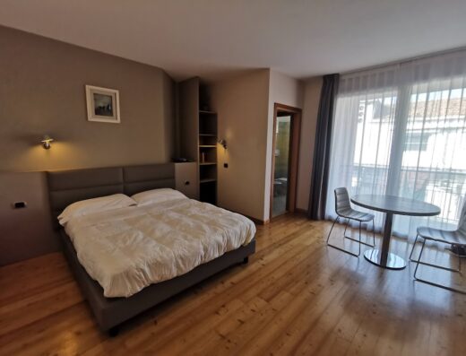 Camera dell'Hotel Rubicone con letto matrimoniale, tavolo rotondo con sedie, pavimento in legno e balcone con tenda.