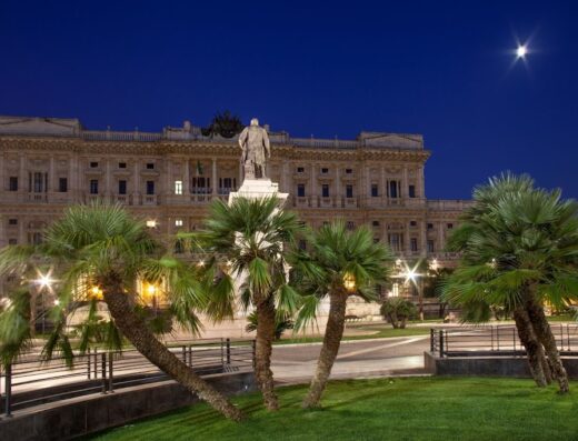 Hotel Sant'Angelo in Rome at night, featuring the Palace of Justice with a statue in front, palm trees, and a bright moon in the sky.