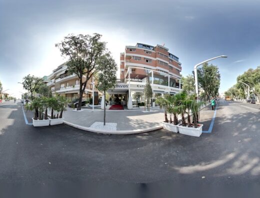 Hotel Savoy vista esterna, ingresso con tappeto rosso, insegna con il nome dell'hotel e caffetteria, palme decorative, traffico cittadino.