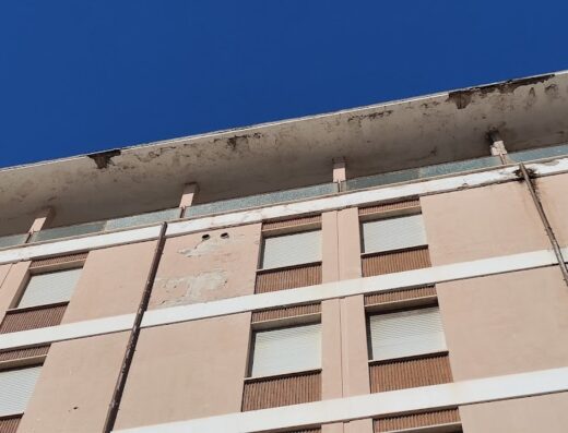 Hotel Sileno: Facciata di un edificio con intonaco deteriorato e finestre chiuse, cielo azzurro sullo sfondo. Dettaglio architettonico.