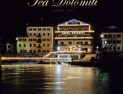 Hotel Tea Dolomiti illuminated at night in Alleghe, Italy, reflecting on the water. A charming three-star hotel in the Dolomites, perfect for a motorcycle travel vacation.