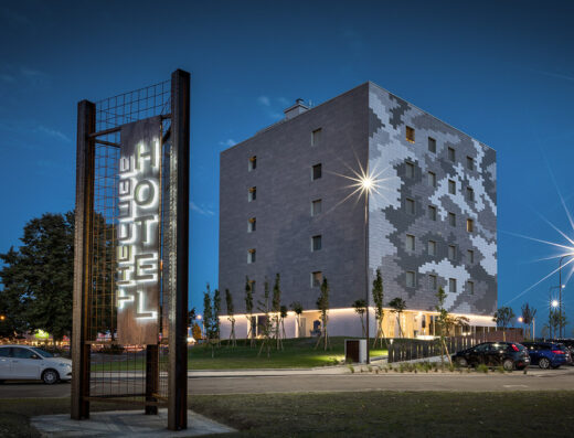 Exterior view of Hotel The Cube at dusk, showcasing its cube-shaped architecture and illuminated sign. Cars are parked nearby.