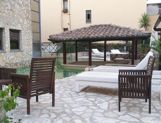 Outdoor lounge area at Hotel the Strand with wooden chairs, white chaise lounges and a covered patio overlooking the pool.