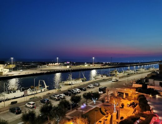 Vista notturna del porto di Fiumicino dal Hotel Tiber Rooftop Wellness, con barche ormeggiate, auto parcheggiate e luci soffuse sul lungomare.