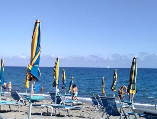 Spiaggia di Hotel Tirreno con ombrelloni chiusi, lettini blu e persone che si godono il mare in una giornata di sole.