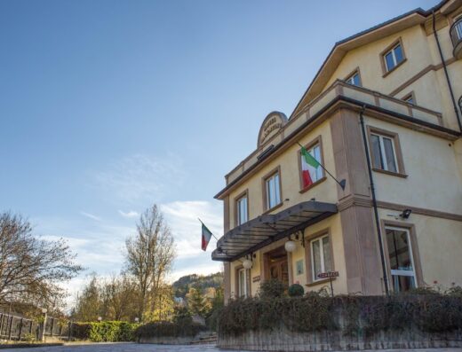 Fotografia dell'Hotel Valentino ad Acqui Terme, con bandiere italiane, architettura elegante e cielo azzurro. Ideale per un soggiorno rilassante e un'esperienza di viaggio indimenticabile.