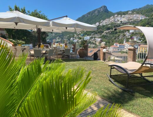 Outdoor dining area at a hotel on the Vietri Coast, Italy, with tables set under umbrellas, a relaxing lounge chair and a scenic view of the mountains and town