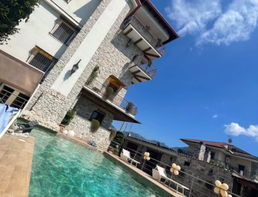 Hotel Villa Clementina: vista della piscina con acqua cristallina, edificio in pietra con balconi, sedie bianche e cielo azzurro.
