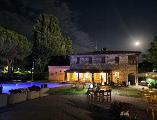 Hotel Villalago Trasimeno at night, featuring a stone building, illuminated pool, outdoor seating, and a full moon in the sky.