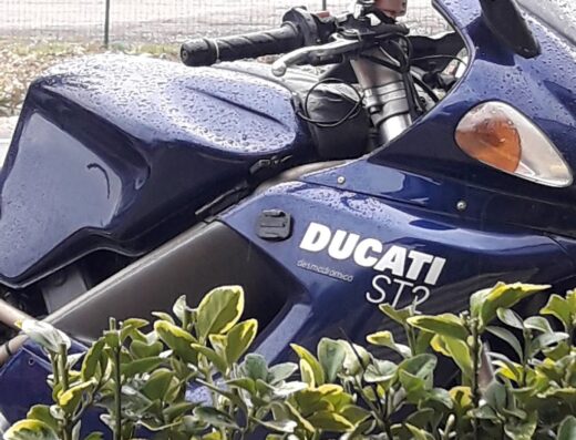 Dettaglio di una moto Ducati ST2 blu coperta di gocce di pioggia, con vegetazione in primo piano e cartello "Autolavaggio a mano" sullo sfondo.