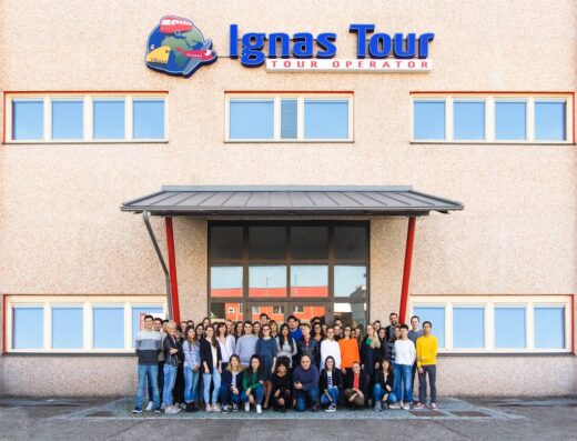 Gruppo di persone di fronte all'edificio Ignas Tour, operatore turistico viaggi e servizi, con logo che raffigura un autobus turistico, un aereo e la terra.