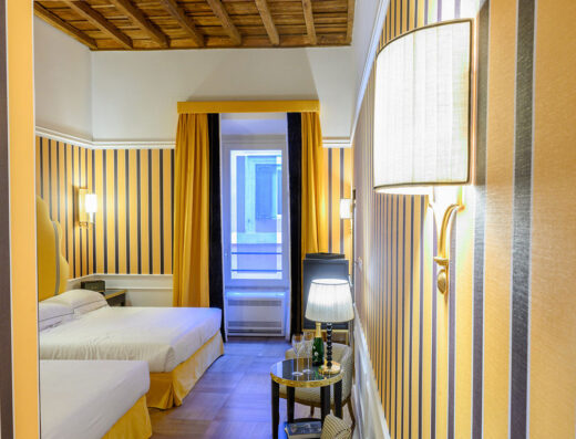 Camera con due letti all'Il Campo Marzio Hotel, arredata con carta da parati a righe verticali gialle e nere, tendaggi gialli, e lampade da parete decorative. Sul tavolino, una bottiglia di champagne e bicchieri.