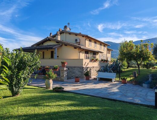 Il Casale di Monica: vista esterna della struttura con giardino curato, dettagli in pietra, vasi di fiori e cielo azzurro.