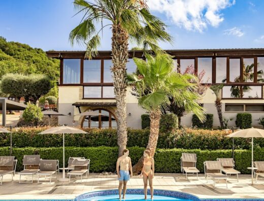 Il Cascinale Country House Ristorante Spa: A couple stands in a pool amidst palm trees and lounge chairs, showcasing the resort's relaxation and spa facilities.