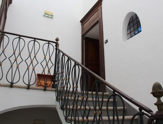 Scala interna in "Il Castello", con ringhiera in ferro battuto decorata e gradini in pietra. Porta in legno aperta e finestra ad arco.