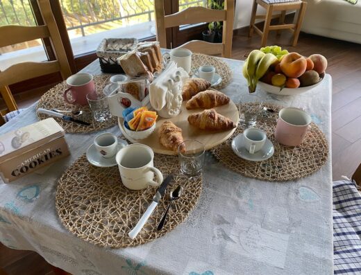 Colazione italiana con croissant freschi, frutta, caffè e biscotti 'American Cookies' su una tavola apparecchiata in un ambiente luminoso.