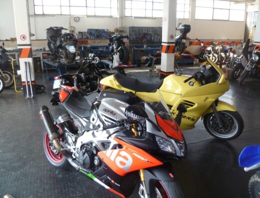 Officina Iliano Racing: una varietà di moto, tra cui un'Aprilia RSV4 e una BMW gialla, in riparazione e manutenzione. Servizi di assistenza moto e marketplace di accessori.