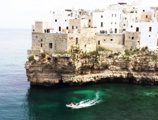 Veduta panoramica di Polignano a Mare, Puglia, con case bianche a picco sulla scogliera e una barca che solca il mare cristallino. Esplora la bellezza dei viaggi in moto e scopri accessori unici nel nostro marketplace.