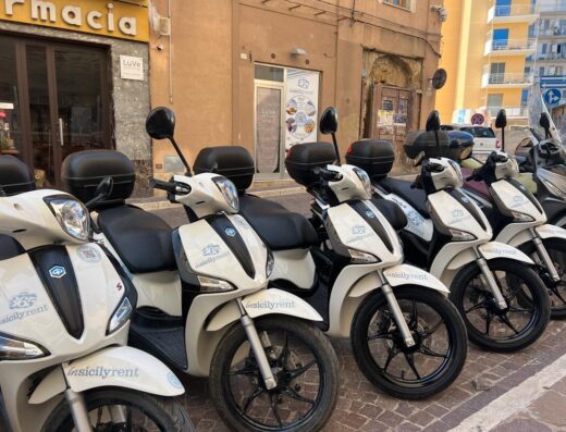 File di scooter a noleggio "In Sicily 365 Travel & Rent City Hub" parcheggiati in strada con bauletto, ideali per esplorare la città.