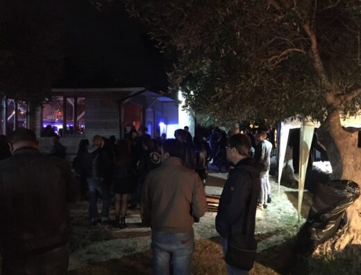 Gruppo di persone riunite di notte vicino a un edificio e un albero, atmosfera potenzialmente legata a un evento motociclistico.