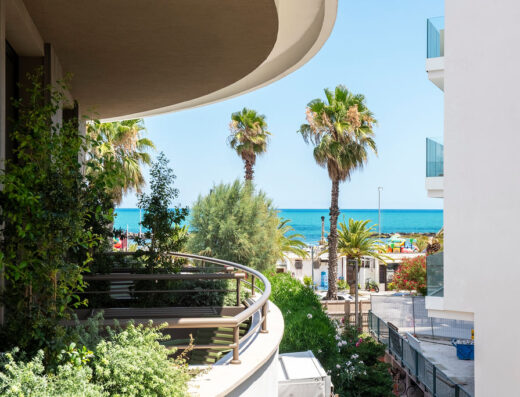 Vista dalla suite internazionale dell'hotel con palme che ondeggiano verso l'oceano turchese, vegetazione lussureggiante e dettagli architettonici moderni.