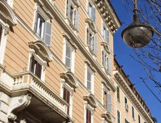 Facciata di un edificio storico a Roma con finestre aperte e chiuse, balconi decorati e un lampione antico sullo sfondo di un cielo azzurro. Architettura tipica romana.