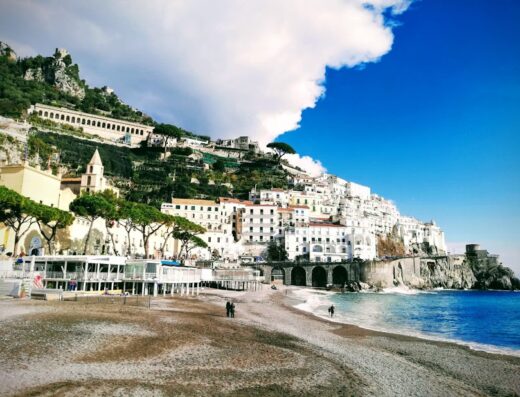Veduta panoramica di Amalfi, Italia, con case bianche arroccate sulla collina, spiaggia di sabbia e mare cristallino. Irno Viaggi di Orlando Rago