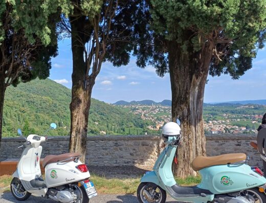 Noleggio Vespa e bici in Italia: due scooter Vespa parcheggiati con casco, sullo sfondo un paesaggio collinare italiano.