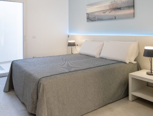 Camera da letto moderna e luminosa con letto matrimoniale, comodini con lampade e accesso a balcone esterno. Arredamento minimalista con quadro a tema marino.