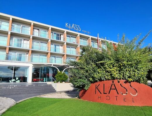 Vista esterna dell'Hotel Klass con insegna in primo piano, architettura moderna, balconi in vetro e vegetazione lussureggiante sotto un cielo azzurro.