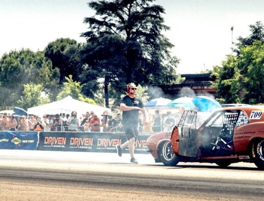 Auto da corsa arancione con numero 709 e sponsor Midland sulla pista, meccanico con cuffie corre vicino, pubblico e gazebo sullo sfondo