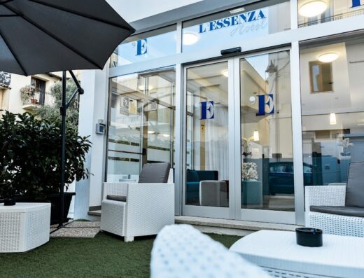 Ingresso dell'Essenza Hotel a Olbia, con arredi esterni in vimini bianco, ombrellone e vetrate con logo. Hotel di design per viaggi e vacanze in Sardegna.