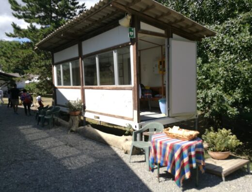 La Bosana agriturismo e centro ippico: struttura prefabbricata con finestre e porta aperta, tavolo con tovaglia colorata e cesto di pane, area esterna con sedie e piante.