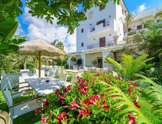 Hotel La Bussola outdoor dining area with straw umbrellas, white tables and chairs, lush tropical plants, and the hotel building in the background.