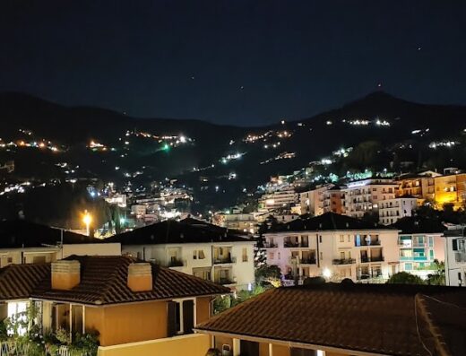 Vista notturna illuminata di una cittadina collinare italiana, con case e luci che si arrampicano sulle montagne sullo sfondo.