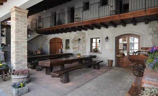 Patio esterno di Là di Muk, con tavoli in legno massiccio, panche rustiche, carretti decorativi con fiori, e balcone in legno, ideale per eventi e relax all'aperto.