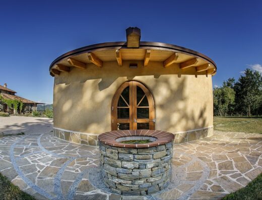 La Fattoria dell'Autosufficienza: Edificio circolare in bioedilizia con tetto in legno e pozzo in pietra, circondato da pavimentazione in pietra naturale e prato verde. Architettura sostenibile e design rurale italiano.