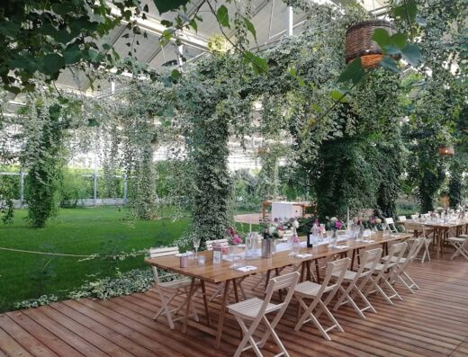 A long wooden table is set with flowers, glasses, and plates, surrounded by white folding chairs, on a wooden deck in a lush greenhouse with hanging vines and greenery.