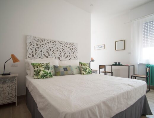 Camera da letto luminosa con testiera decorata, cuscini a tema tropicale, scrivania con sedie e termosifone verde acqua, per un soggiorno confortevole a La Maison Extravagante.