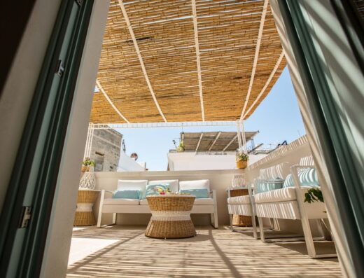 Terrazza panoramica a La Naca sul Porto, Puglia: arredamento esterno con divani bianchi, cuscini azzurri, tavolino in vimini e tettoia di canne, ideale per rilassarsi e godersi la vista sul mare.