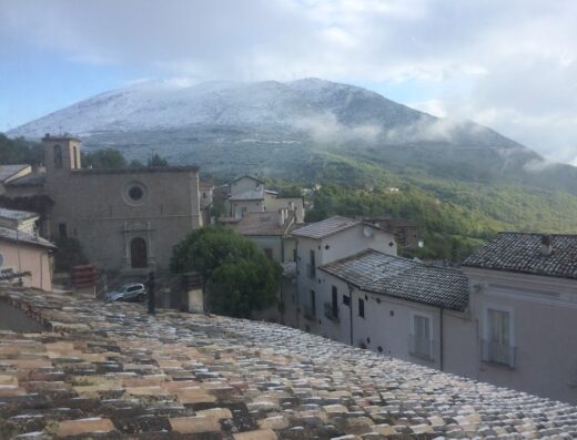 Scenic view of a mountain partially covered in snow, seen from a rooftop in La Porta dei Parchi, with a church in the foreground and houses with tiled roofs.