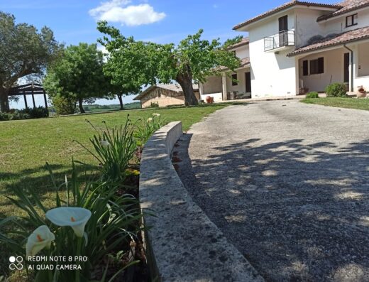 Bella casa di campagna con vialetto in cemento, giardino curato e cielo azzurro, ideale per un viaggio in moto alla scoperta della Toscana.