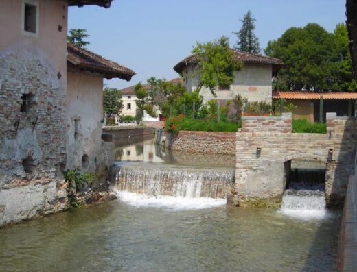 Scorcio pittoresco de La Torre del Falco, con un canale d'acqua che scorre tra edifici storici in pietra e mattoni, creando una cascata suggestiva. Un'oasi di tranquillità e storia.